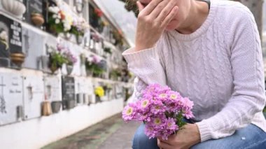  Mezarlıkta elinde taze çiçeklerle mezar duvarının yanında oturmuş ağlayan ve kafaya dokunan günlük kıyafetlerle yas tutan bir kadın.