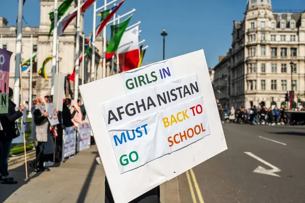 Protesting people with posters while standing on London street with banner containing inscription girls in Afghanistan must go back to school on pavement against buildings and blue sky