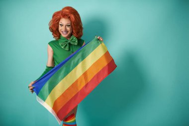 Side view of transgender male model in stylish feminine bodysuit with LGBT rainbow flag looking at camera while standing in studio against blue background
