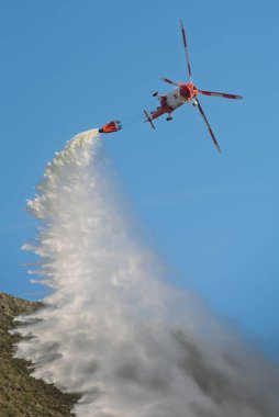 Uçan itfaiye helikopterinden, bulutsuz mavi gökyüzüne karşı, güneşli bir havada, kayalık arazide, Bambi kovasından su salıyor.