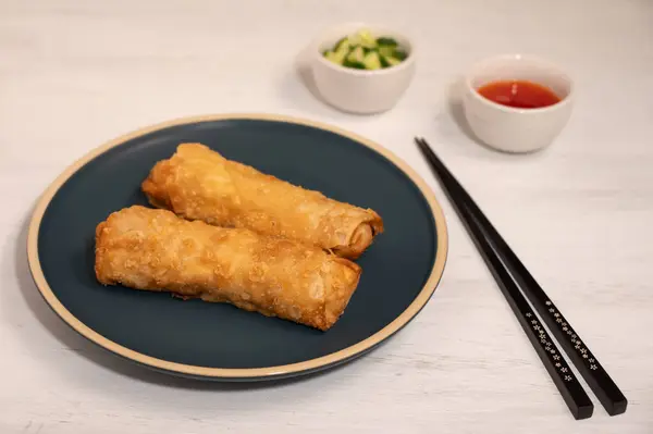 Two spring rolls on a dark blue plate alongside a pair of chopsticks and two small dishes of chilli sauce and cucumbers.