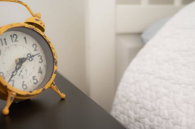 Beyaz yorganı olan beyaz bir yatağın yanındaki siyah komodinin üzerindeki sarı alarm saati..