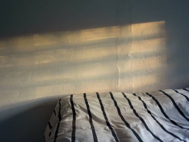 Pencere çerçevesinin gölgesi ve gün batımının ışığı siyah çizgili beyaz yorganla kaplı dağınık bir yatağın üzerinde..