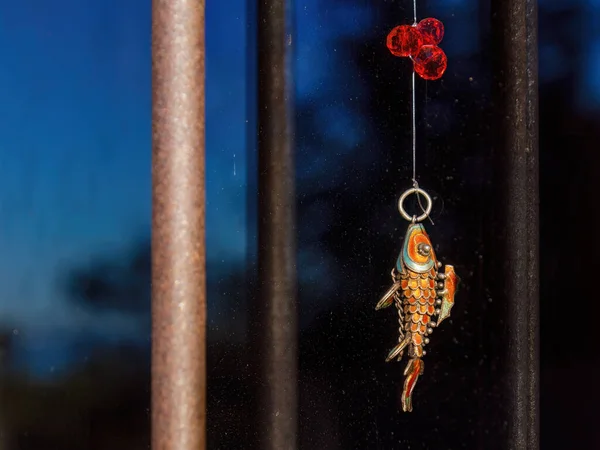 A colorful metallic fish with three red plastics beans, hanging on a window and illuminated by the light of the sunrise, in a house in central Colombia.
