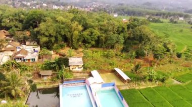 Güneşli bir günde pirinç tarlaları manzaralı bir tatil köyünden özel havuz. Bandung, Endonezya 'nın dışındaki pirinç tarlalarının kenarındaki özel bir havuzun insansız hava aracı görüntüsü. Yüksek Kalite 4K Drone Görüntüsü