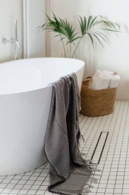 Klasik küveti olan beyaz bir otel. Banyodaki fayanslı zeminde su gideri var. Hasır sepetin içinde havlu ve küvetin üzerine atılmış kumaş. Minimalist dairede modern iç tasarım kavramı