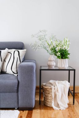 İskandinav iç dekorasyonu, yastıklı gri kanepe, cam sehpada seramik vazoda çiçekler ve ahşap zeminde ekose sepet.