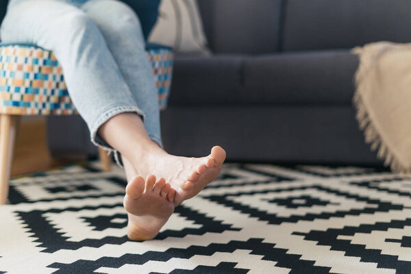 cropped shot of female with barefoot legs on floor carpet relaxing on comfort armchair near couch in living room at cozy apartment