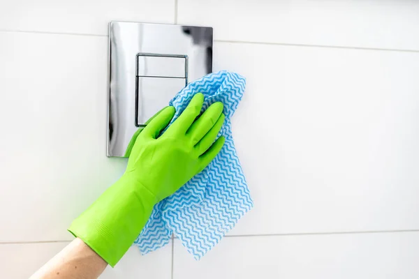 female hand in green rubber glove with fabric rag wash wc flush button built in wall with white tile in modern apartment bathroom, cropped shot, cleaning service concept