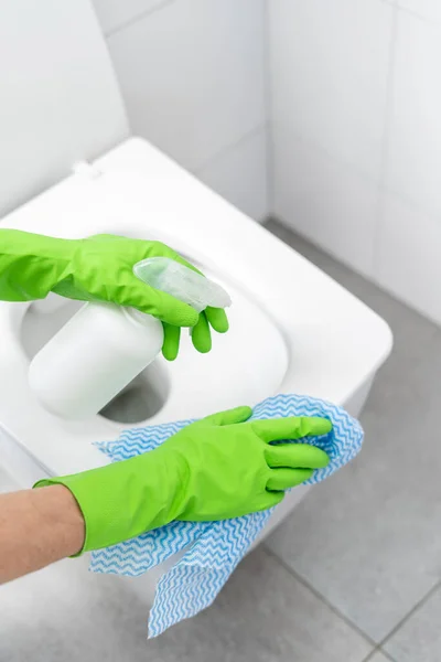 female hand in green rubber glove washing wc lid with fabric rag and disinfect with spray detergent in apartment bathroom, cropped shot, cleaning service concept