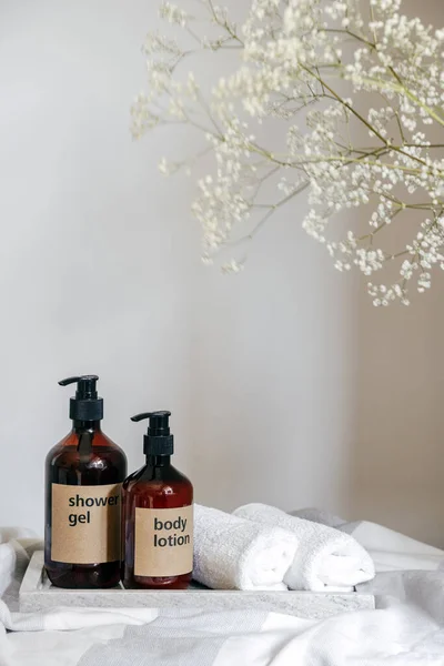 composition of shower gel and body lotion in brown plastic bottles with dispenser, white clean bath towel folded on marble tray and flower at home bathroom, personal care concept