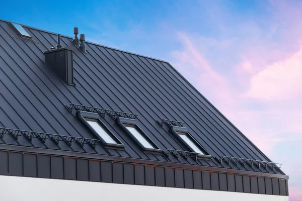 House with metal galvanized coating, chimney pipe, mansard windows and snow holding cornice on roof slope against sunset sky. Element of modern residential building with zinc rooftop sheets
