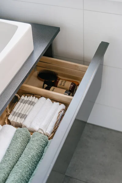 open drawer with fresh terry towels tidy folded in wicker basket and wooden box with hair brush and cosmetic bottles, organized storage at modern house