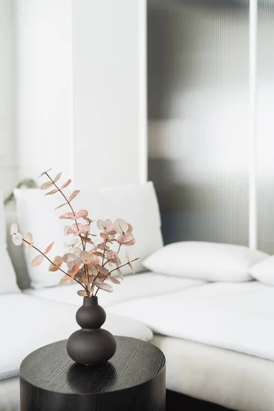 eucalyptus branch in ceramic vase on wooden round side table and couch with pillows on background in apartment with scandinavian interior