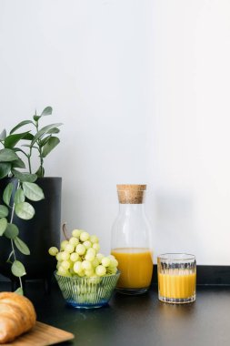 Mutfağın içindeki detaylar, taze meyve püresi ya da şişedeki mantar kapaklı sağlıklı içecek, ahşap tepside kruvasan ve mutfaktaki siyah tezgahta beyaz duvarlı saksı bitkisi.