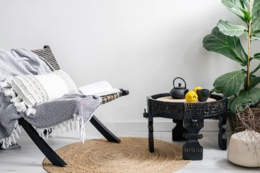 Siyah bir koltukta ekoseli retro oturma odasının iç tasarımı, Çin çayı için çaydanlığı olan ahşap yan masa, şık ev dekorasyonu ve beyaz fotokopi duvarına karşı yeşil ev bitkileri.