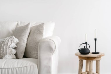 Siyah çaydanlık ve mum tutacağı ile ahşap masaya yakın dekoratif yastıklı rahat beyaz kanepe sahnesi, fotokopi alanı arka planına karşı. İskandinav tarzında oturma odası