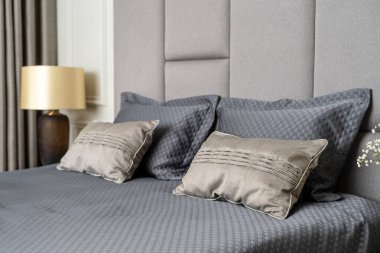 İç döşeme, yumuşak yastıklar ve rahat yatak başlığıyla gri battaniye örtülü yatak odası, lüks yatak odasındaki beyaz duvarın yanında şık bir lamba.