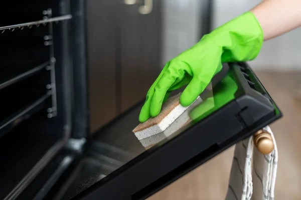 cropped shot of female hand in rubber glove with sponge scrubber cleaning open door of built in oven at kitchen, housework concept