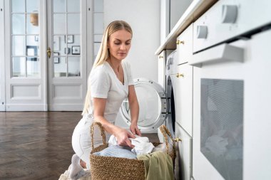 Kadın hasır çamaşır sepetinden kirli çamaşırları alır ve mutfaktaki otomatik çamaşır makinesine koyar. Modern aletler, ev idaresi ve ev işi kavramı