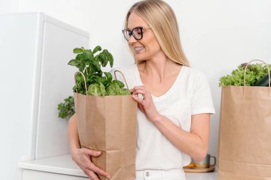 Market alışverişi ve teslimat konsepti. Gülümseyen bir kadının elinde taze yeşil salata yaprakları, marul ve fesleğen bitkileriyle birlikte beyaz ev mutfağında duran çevre dostu bir alışveriş çantası var.