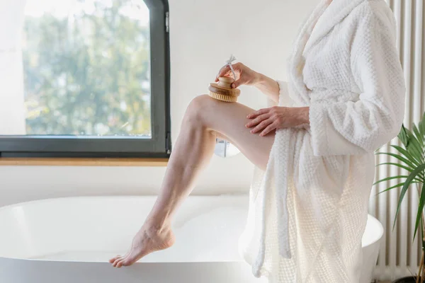 Profile view of woman brushing body in circular motions or light strokes to provide exfoliation and rejuvenating boost of energy. Female wearing at white bathrobe using wooden massager for legs