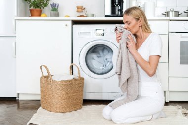 advertising of fabric softener, delicate laundry cycle. female housewife holding and smelling soft fresh clothes after washing, sitting near wicker basket and automatic washing machine
