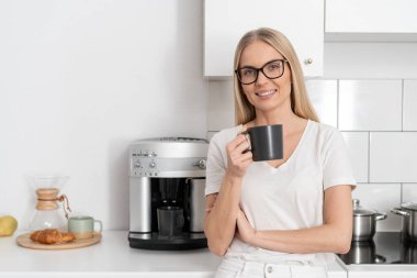 happy woman with cup of beverage looking at camera. female enjoying espresso, making drink with coffee machine while she standing at kitchen. light interior and modern household appliances