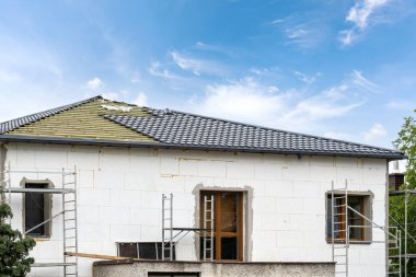 view of unfinished building from concrete blocks and rooftop with wooden truss planks and dark grey tile layer outdoors on blue sky background