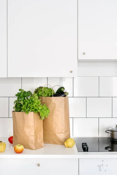 grocery purchase and delivery concept. full recycled craft or paper bags with fresh vegetables, salad leaves and green herbs on worktop at home kitchen with white tile