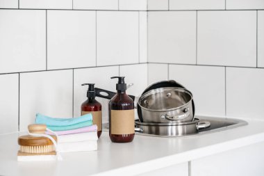 microfiber cloth, soap and dishwashing liquid in dispenser bottles with copy space, eco scrubber sponge and bamboo brush near sink with dirty saucepans at kitchen, zero waste concept