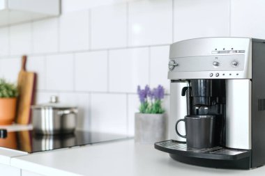 selective focus on automatic espresso machine with dark mug on white countertop at stylish kitchen with household appliances and kitchenware on blurred background