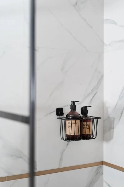 compact black metal rack organizer with shower gel and body lotion in brown bottles on wall with marble tile in bathroom with modern interior