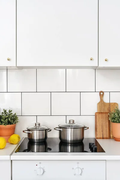 detail in kitchen with light interior, white cupboard furniture, tile on wall, modern electrical hob, stainless steel saucepan with lid and plant in pot