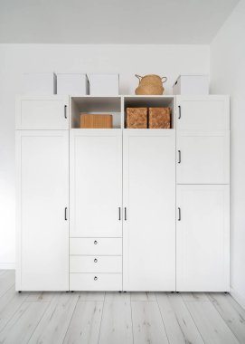 front view of modern white wardrobe with closed doors, drawers and wicker baskets on shelves in dressing room with light interior, organized storage concept