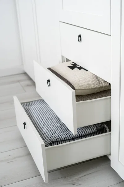 side view of open drawers in wooden modern wardrobe with pillows, folded blanket and bedclothes inside in bedroom, organized storage concept