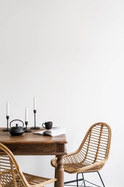 Çin çaydanlığı, seramik bardak, metal şamdanlar, ahşap masa üzerinde açık kitap ve beyaz duvar arkasında bambu rattan sandalyeler.