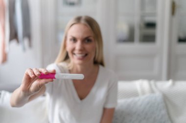 Hamilelik testinde kamera önünde pozitif sonuç veren neşeli bir kadın ve oturma odasında kanepede otururken gülümseyen bir kadın.