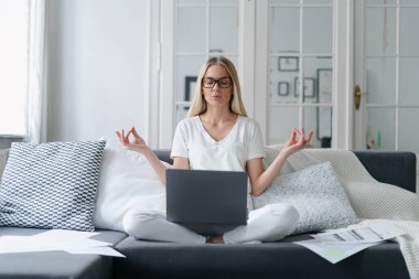 Gözlüklü kadın meditasyon yaparken dizüstü bilgisayarda, oturma odasında rahat koltukta otururken sakin bir konsept, stres atma, uzaktan çalışma.