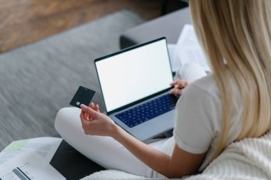 Kadın elinde kredi kartı ve dizüstü bilgisayarla kanepeye oturmuş, internet bankacılığı kavramları ve ani para transferleri yaparken online alışveriş yapıyor.