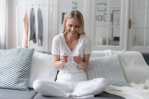 stock image joyful female looking on positive result at pregnancy test and smiling while sitting on sofa in living room, maternity concept