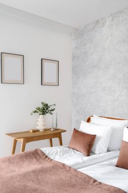Evin içinde şık bir yatak odası, yatak örtülü rahat bir yatak, yumuşak yastıklar ve kahverengi tekstil örtüsü, mum ve vazolu ahşap masa, duvarda sahte resimler.