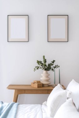 Otel odasının içi rahat, ahşap masa üzerinde mumlar, seramik vazoda okaliptüs dalı, mücevher kutusu ve yatak döşekleriyle çerçeveli resimler.