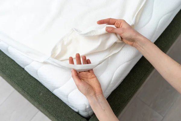 cropped shot of female hands putting cotton bed sheet with elastic band on mattress corner in bedroom, protection cover, bedclothes change concept