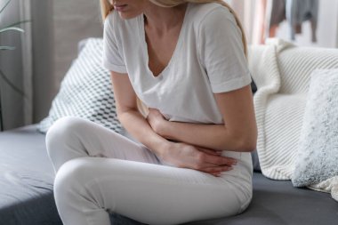 cropped shot of young female with stomach ache feeling unwell, holding hands on belly and sitting on couch in living room at home, girl with indigestion clipart