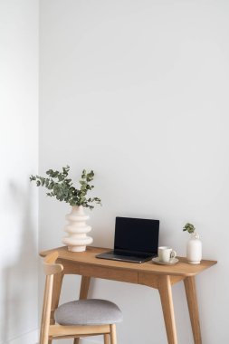 Laptop, okaliptüs dallı seramik vazo ve beyaz duvarlı modern apartman dairesinin rahat sandalyesinin yanındaki ahşap masada bir fincan kahve.