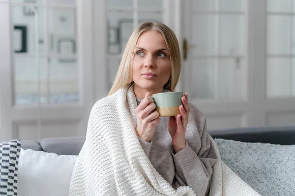 young female with influenza sitting on sofa, covered with warm knitted blanket, holding cup of tea and feeling recovery and relief, health care concept