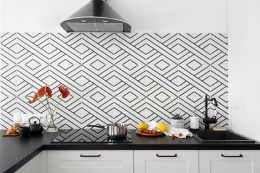 Şık bir iç mimarisi olan mutfak, siyah lavabo ve musluk, elektrikli ocakta tencere, domatesli tabak, saksı bitkisi, cam vazoda çiçekler, tezgahta fincanı olan çaydanlık ve duvarda desenli fayans.