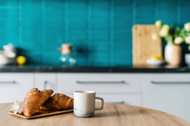 Bambu tabağındaki taze kruvasanlara yakın çekim. Süslü çiçek ve ahşap yemek masasında bir fincan kahve. Mutfak arka planında bulanık kahvaltı kavramı.