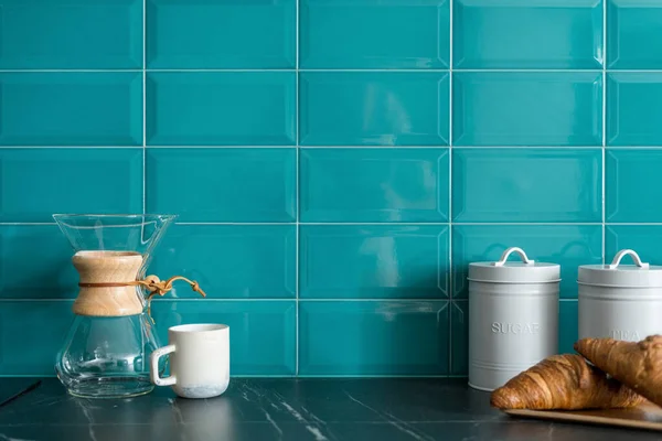 glass coffee brewing equipment, ceramic cup, fresh croissants on wooden plate and food storage containers with sugar and tea on countertop at stylish kitchen with blue tile on wall